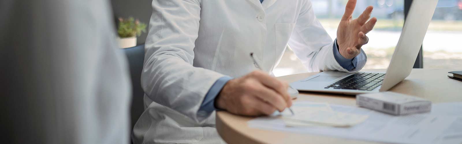 Young male doctor talking to a patient in an office working on laptop